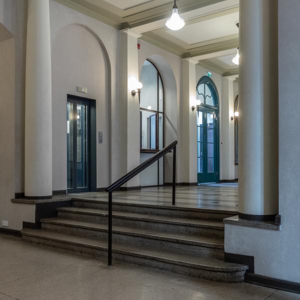 Hallrum med fyra trappsteg till hallens övre plan och ett räcke i mitten av trappan. I övre ändan av trappan finns hiss.