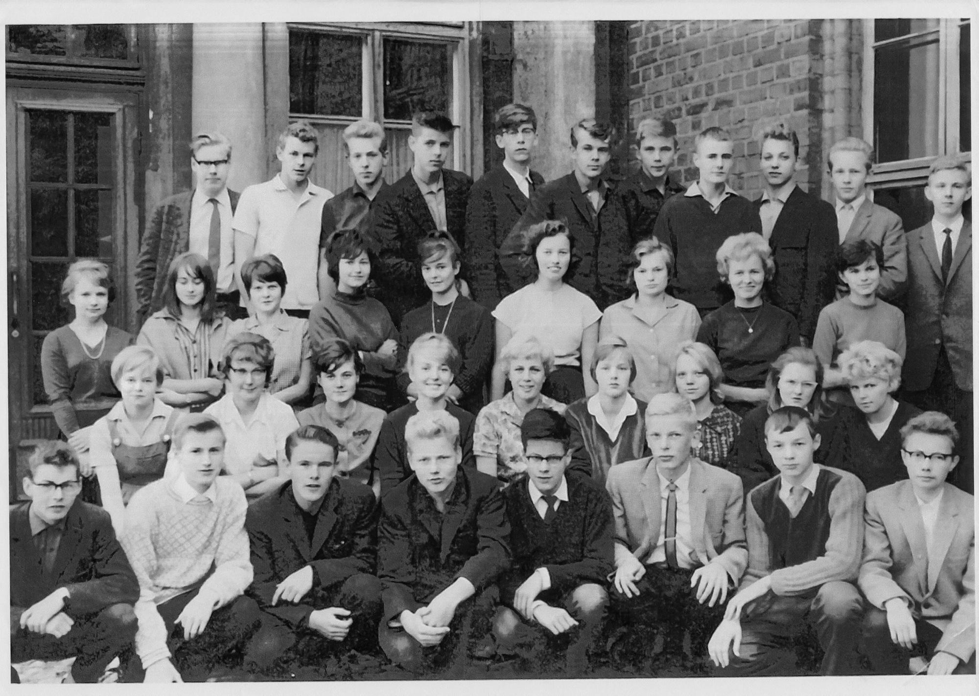 Mustavalkoinen vanha luokkakuva, jossa tyttöja ja poikia riveittäin rakennuksen edustalla.