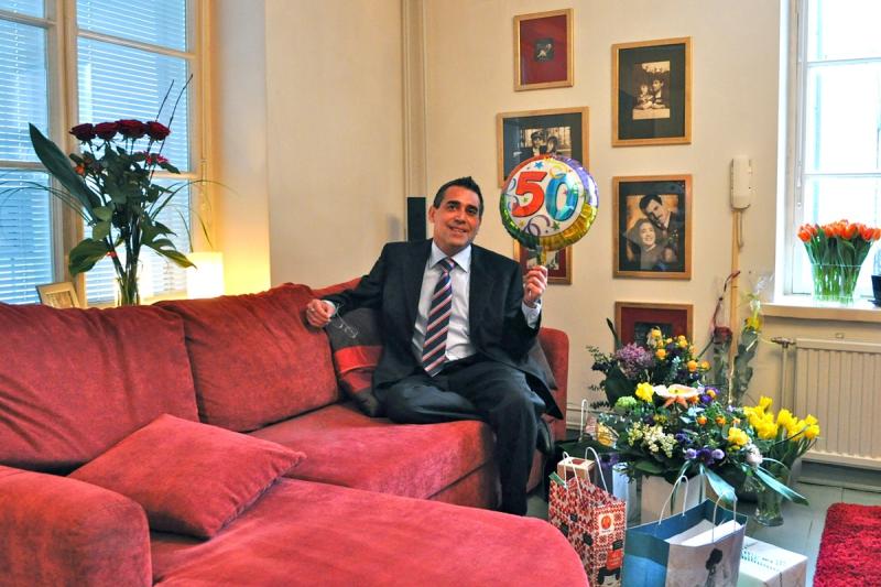 Miesoletettu henkilö sohvalla, kädessään ilmapallo ja vieressä paljon kukkia.