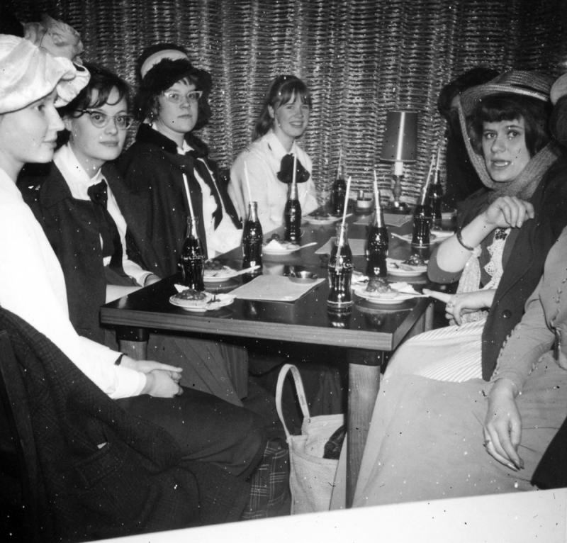 Vanha kuva, jossa nuoria naisia hienosti pukeutuneena pöydän äärellä colapullot edessään.